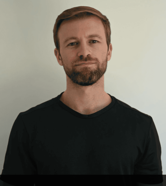 Bearded man with short brown hair wearing a black t-shirt against a plain white background.