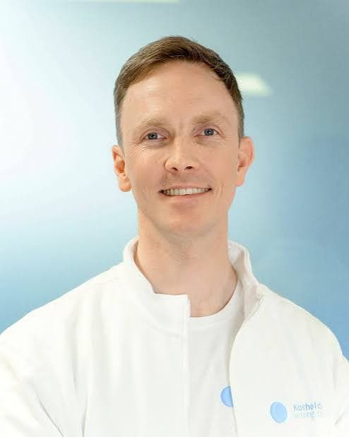 Smiling man with short brown hair wearing a white medical coat against a blue background.